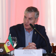 A imagem mostra um participante sentado à mesa de reunião, posicionado diante de uma bandeira do Rio Grande do Sul e de uma cuia com chimarrão colocadas sobre a mesa. Há um microfone direcionado para ele, além de copos e materiais de apoio. O fundo tem cortinas claras iluminadas por luz rosada.
