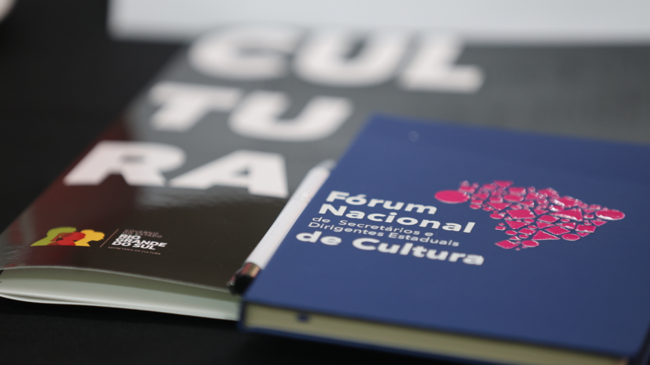 A imagem mostra dois materiais gr&aacute;ficos sobre uma mesa preta: um caderno azul com o logotipo do F&oacute;rum Nacional de Secret&aacute;rios e Dirigentes Estaduais de Cultura e um material impresso com a palavra &ldquo;CULTURA&rdquo; em letras grandes e brancas sobre fundo escuro. Ambos est&atilde;o dispostos lado a lado, destacando a identidade visual do evento.
