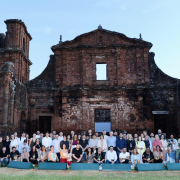 A imagem mostra um grande grupo reunido diante da fachada de pedra das ruínas missioneiras, posando para uma foto coletiva sobre uma área de gramado. A construção histórica aparece imponente ao fundo, com sua textura avermelhada e suas aberturas características. O céu azul completa o cenário.
