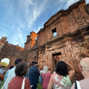 A imagem mostra visitantes reunidos diante da fachada de pedra avermelhada das ruínas de uma antiga construção missioneira. O grupo observa a estrutura histórica enquanto o céu azul com nuvens suaves se estende acima do prédio. A fachada tem grandes aberturas e paredes marcadas pelo desgaste do tempo. O gramado verde ocupa a parte inferior da cena.

