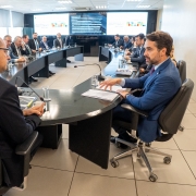 Sala de reuni&otilde;es moderna com uma mesa grande em formato de U. Cerca de dez homens de terno est&atilde;o sentados ao redor, participando de uma reuni&atilde;o formal. Em primeiro plano, dois homens conversam; um deles gesticula enquanto fala, com pap&eacute;is &agrave; sua frente. Sobre a mesa h&aacute; copos de &aacute;gua, x&iacute;caras de caf&eacute;, microfones e computadores. Ao fundo, grandes telas mostram uma apresenta&ccedil;&atilde;o com gr&aacute;ficos coloridos e texto. A luz entra pelas janelas laterais, iluminando o ambiente corporativo.
