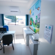 Consult&oacute;rio infantil com decora&ccedil;&atilde;o tem&aacute;tica colorida em uma das paredes, incluindo ilustra&ccedil;&otilde;es de animais e natureza. O espa&ccedil;o possui maca, mesa com computador, cadeiras e janela ampla com cortinas curtas, al&eacute;m de ilumina&ccedil;&atilde;o branca no teto.