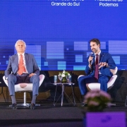 Tr&ecirc;s pessoas est&atilde;o sentadas em poltronas no palco de um evento. Um homem de terno cinza e gravata vermelha est&aacute; &agrave; esquerda, outro homem de terno azul fala ao microfone no centro, e uma mulher com roupa colorida est&aacute; &agrave; direita. Atr&aacute;s deles h&aacute; um grande painel azul com textos.