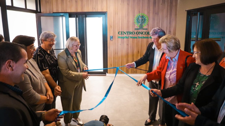 Esta &eacute; uma descri&ccedil;&atilde;o da imagem para fins de acessibilidade:

A fotografia registra o momento inaugural do novo Centro Oncol&oacute;gico do Hospital Nossa Senhora da Oliveira, em Vacaria. Um grupo de sete pessoas participa do ato simb&oacute;lico de desatamento da fita.

Elementos Principais
O Ato: No centro da imagem, as pessoas est&atilde;o posicionadas em duas alas (tr&ecirc;s &agrave; esquerda e quatro &agrave; direita), segurando e puxando as pontas de uma fita de cetim azul. O movimento indica o exato momento em que o la&ccedil;o &eacute; desfeito.

As Pessoas: O grupo &eacute; composto majoritariamente por mulheres.

&Agrave; esquerda, destaca-se uma mulher de blazer bege e &oacute;culos, sorrindo enquanto segura a fita.

&Agrave; direita, uma mulher veste um blazer vermelho vibrante sobre uma camisa listrada, tamb&eacute;m participando ativamente do desenlace.

Os demais presentes vestem trajes formais e casuais, exibindo express&otilde;es de satisfa&ccedil;&atilde;o e celebra&ccedil;&atilde;o.

O Cen&aacute;rio: Ao fundo, em uma parede com revestimento amadeirado, est&aacute; fixado o letreiro do local: "CENTRO ONCOL&Oacute;GICO - Hospital Nossa Senhora da..." (o restante do nome est&aacute; parcialmente encoberto). Acima do texto, h&aacute; um logotipo verde em formato de &aacute;rvore estilizada.

Detalhes e Ambiente
Arquitetura: O ambiente &eacute; moderno, com piso de porcelanato cinza claro e portas de vidro com esquadrias pretas ao fundo, que trazem claridade ao espa&ccedil;o.

Composi&ccedil;&atilde;o: A foto captura o grupo em um plano m&eacute;dio, focando na intera&ccedil;&atilde;o entre as pessoas e a fita. No canto inferior central, nota-se o topo da cabe&ccedil;a de um fot&oacute;grafo e sua lente, indicando a cobertura oficial do evento.

Ilumina&ccedil;&atilde;o: A luz &eacute; interna e uniforme, proveniente de lumin&aacute;rias de LED embutidas no teto, criando um ambiente acolhedor e profissional.