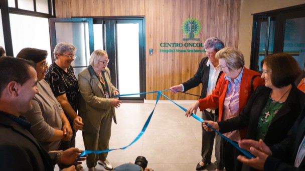 Esta &eacute; uma descri&ccedil;&atilde;o da imagem para fins de acessibilidade:

A fotografia registra o momento inaugural do novo Centro Oncol&oacute;gico do Hospital Nossa Senhora da Oliveira, em Vacaria. Um grupo de sete pessoas participa do ato simb&oacute;lico de desatamento da fita.

Elementos Principais
O Ato: No centro da imagem, as pessoas est&atilde;o posicionadas em duas alas (tr&ecirc;s &agrave; esquerda e quatro &agrave; direita), segurando e puxando as pontas de uma fita de cetim azul. O movimento indica o exato momento em que o la&ccedil;o &eacute; desfeito.

As Pessoas: O grupo &eacute; composto majoritariamente por mulheres.

&Agrave; esquerda, destaca-se uma mulher de blazer bege e &oacute;culos, sorrindo enquanto segura a fita.

&Agrave; direita, uma mulher veste um blazer vermelho vibrante sobre uma camisa listrada, tamb&eacute;m participando ativamente do desenlace.

Os demais presentes vestem trajes formais e casuais, exibindo express&otilde;es de satisfa&ccedil;&atilde;o e celebra&ccedil;&atilde;o.

O Cen&aacute;rio: Ao fundo, em uma parede com revestimento amadeirado, est&aacute; fixado o letreiro do local: "CENTRO ONCOL&Oacute;GICO - Hospital Nossa Senhora da..." (o restante do nome est&aacute; parcialmente encoberto). Acima do texto, h&aacute; um logotipo verde em formato de &aacute;rvore estilizada.

Detalhes e Ambiente
Arquitetura: O ambiente &eacute; moderno, com piso de porcelanato cinza claro e portas de vidro com esquadrias pretas ao fundo, que trazem claridade ao espa&ccedil;o.

Composi&ccedil;&atilde;o: A foto captura o grupo em um plano m&eacute;dio, focando na intera&ccedil;&atilde;o entre as pessoas e a fita. No canto inferior central, nota-se o topo da cabe&ccedil;a de um fot&oacute;grafo e sua lente, indicando a cobertura oficial do evento.

Ilumina&ccedil;&atilde;o: A luz &eacute; interna e uniforme, proveniente de lumin&aacute;rias de LED embutidas no teto, criando um ambiente acolhedor e profissional.