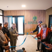 Esta &eacute; uma descri&ccedil;&atilde;o da imagem para fins de acessibilidade:

A fotografia registra o momento inaugural do novo Centro Oncol&oacute;gico do Hospital Nossa Senhora da Oliveira, em Vacaria. Um grupo de sete pessoas participa do ato simb&oacute;lico de desatamento da fita.

Elementos Principais
O Ato: No centro da imagem, as pessoas est&atilde;o posicionadas em duas alas (tr&ecirc;s &agrave; esquerda e quatro &agrave; direita), segurando e puxando as pontas de uma fita de cetim azul. O movimento indica o exato momento em que o la&ccedil;o &eacute; desfeito.

As Pessoas: O grupo &eacute; composto majoritariamente por mulheres.

&Agrave; esquerda, destaca-se uma mulher de blazer bege e &oacute;culos, sorrindo enquanto segura a fita.

&Agrave; direita, uma mulher veste um blazer vermelho vibrante sobre uma camisa listrada, tamb&eacute;m participando ativamente do desenlace.

Os demais presentes vestem trajes formais e casuais, exibindo express&otilde;es de satisfa&ccedil;&atilde;o e celebra&ccedil;&atilde;o.

O Cen&aacute;rio: Ao fundo, em uma parede com revestimento amadeirado, est&aacute; fixado o letreiro do local: "CENTRO ONCOL&Oacute;GICO - Hospital Nossa Senhora da..." (o restante do nome est&aacute; parcialmente encoberto). Acima do texto, h&aacute; um logotipo verde em formato de &aacute;rvore estilizada.

Detalhes e Ambiente
Arquitetura: O ambiente &eacute; moderno, com piso de porcelanato cinza claro e portas de vidro com esquadrias pretas ao fundo, que trazem claridade ao espa&ccedil;o.

Composi&ccedil;&atilde;o: A foto captura o grupo em um plano m&eacute;dio, focando na intera&ccedil;&atilde;o entre as pessoas e a fita. No canto inferior central, nota-se o topo da cabe&ccedil;a de um fot&oacute;grafo e sua lente, indicando a cobertura oficial do evento.

Ilumina&ccedil;&atilde;o: A luz &eacute; interna e uniforme, proveniente de lumin&aacute;rias de LED embutidas no teto, criando um ambiente acolhedor e profissional.