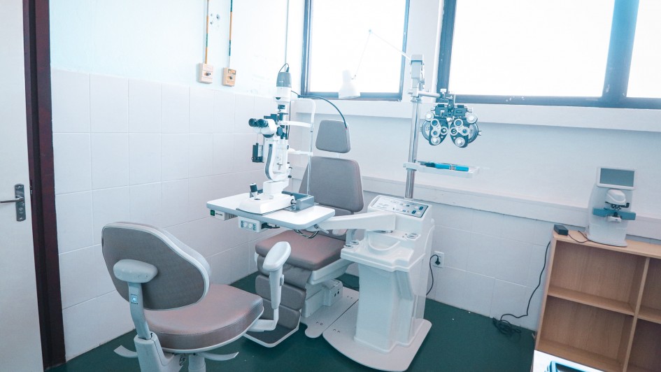 A imagem exibe um consult&oacute;rio oftalmol&oacute;gico equipado com cadeira de exame, l&acirc;mpada de fenda, refrator e mesa ajust&aacute;vel. H&aacute; duas janelas amplas que iluminam a sala, al&eacute;m de ar-condicionado instalado acima delas. 