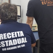 Dois homens de costas, um sentado e outro em p&eacute;, vestindo camisas com a inscri&atilde;o Receita Estadual Rio Grande do Sul e Opera&ccedil;&otilde;es, analisam computadores