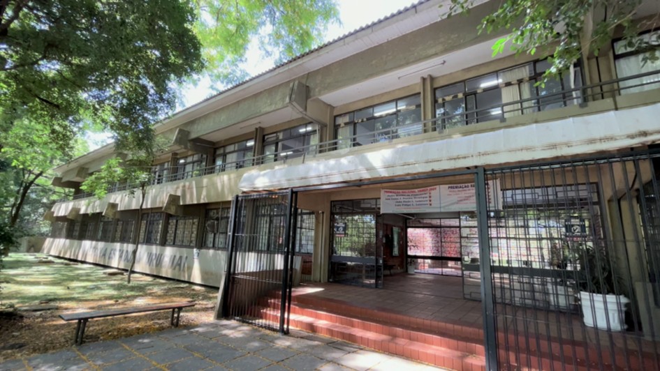 Foto mostra fachada da Escola Uruguai, pr&eacute;dio de dois andares em linha reta. Na frente h&aacute; arvores e um espa&ccedil;o com sombra. 