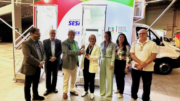 Um grupo de pessoas est&aacute; alinhado em frente a uma unidade m&oacute;vel de sa&uacute;de. Todas est&atilde;o posicionadas sob o toldo lateral e diante das escadas de acesso. A identidade visual do SESI, FIERGS e governo estadual aparece ao fundo, destacada pela ilumina&ccedil;&atilde;o da estrutura.
