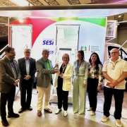 Um grupo de pessoas est&aacute; alinhado em frente a uma unidade m&oacute;vel de sa&uacute;de. Todas est&atilde;o posicionadas sob o toldo lateral e diante das escadas de acesso. A identidade visual do SESI, FIERGS e governo estadual aparece ao fundo, destacada pela ilumina&ccedil;&atilde;o da estrutura.
