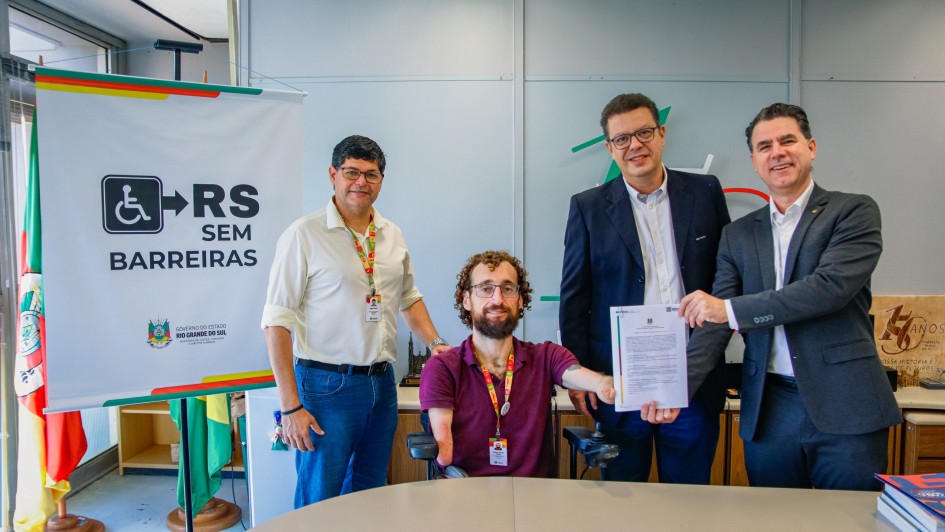 A fotografia registra o momento formal de assinatura de um contrato entre a Secretaria de Justi&ccedil;a, Cidadania e Direitos Humanos (SJCDH) e a General Motors (GM), focado em acessibilidade.

Elementos Principais
Os Participantes: Quatro homens est&atilde;o posicionados atr&aacute;s de uma mesa de escrit&oacute;rio clara.

No centro, um homem com barba e cabelos cacheados, que utiliza uma cadeira de rodas motorizada, segura o contrato assinado junto com o representante &agrave; sua direita.

&Agrave; direita, um homem de terno cinza e sorriso aberto segura a outra extremidade do documento.

&Agrave; esquerda, um homem de camisa branca e cal&ccedil;a jeans, e outro de terno azul marinho e &oacute;culos ao centro-direita, completam o grupo.

O Documento: Um papel oficial com o logotipo do Governo do Estado &eacute; exibido para a c&acirc;mera, simbolizando a formaliza&ccedil;&atilde;o da parceria.

Sinaliza&ccedil;&atilde;o Institucional: &Agrave; esquerda do grupo, h&aacute; um banner branco com a identidade visual da campanha "RS SEM BARREIRAS". O logotipo mostra o s&iacute;mbolo internacional de acesso (cadeira de rodas) seguido de uma seta e o texto em letras pretas garrafais. Abaixo, consta o bras&atilde;o do Governo do Estado do Rio Grande do Sul.

Detalhes e Ambiente
Cen&aacute;rio: O evento ocorre em um escrit&oacute;rio moderno com paredes de divis&oacute;rias cinzas. Ao fundo, &agrave; direita, h&aacute; uma placa decorativa de madeira comemorativa de "70 Anos". &Agrave; esquerda, &eacute; poss&iacute;vel ver parte da bandeira do Rio Grande do Sul.

Composi&ccedil;&atilde;o: A foto &eacute; um plano m&eacute;dio, capturando todos os integrantes da cintura para cima (ou por inteiro, no caso do cadeirante), transmitindo uma atmosfera de coopera&ccedil;&atilde;o e inclus&atilde;o.

Ilumina&ccedil;&atilde;o: A luz &eacute; clara e uniforme, t&iacute;pica de ambientes corporativos, destacando a clareza das informa&ccedil;&otilde;es no banner e no contrato.