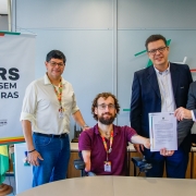 A fotografia registra o momento formal de assinatura de um contrato entre a Secretaria de Justi&ccedil;a, Cidadania e Direitos Humanos (SJCDH) e a General Motors (GM), focado em acessibilidade.

Elementos Principais
Os Participantes: Quatro homens est&atilde;o posicionados atr&aacute;s de uma mesa de escrit&oacute;rio clara.

No centro, um homem com barba e cabelos cacheados, que utiliza uma cadeira de rodas motorizada, segura o contrato assinado junto com o representante &agrave; sua direita.

&Agrave; direita, um homem de terno cinza e sorriso aberto segura a outra extremidade do documento.

&Agrave; esquerda, um homem de camisa branca e cal&ccedil;a jeans, e outro de terno azul marinho e &oacute;culos ao centro-direita, completam o grupo.

O Documento: Um papel oficial com o logotipo do Governo do Estado &eacute; exibido para a c&acirc;mera, simbolizando a formaliza&ccedil;&atilde;o da parceria.

Sinaliza&ccedil;&atilde;o Institucional: &Agrave; esquerda do grupo, h&aacute; um banner branco com a identidade visual da campanha "RS SEM BARREIRAS". O logotipo mostra o s&iacute;mbolo internacional de acesso (cadeira de rodas) seguido de uma seta e o texto em letras pretas garrafais. Abaixo, consta o bras&atilde;o do Governo do Estado do Rio Grande do Sul.

Detalhes e Ambiente
Cen&aacute;rio: O evento ocorre em um escrit&oacute;rio moderno com paredes de divis&oacute;rias cinzas. Ao fundo, &agrave; direita, h&aacute; uma placa decorativa de madeira comemorativa de "70 Anos". &Agrave; esquerda, &eacute; poss&iacute;vel ver parte da bandeira do Rio Grande do Sul.

Composi&ccedil;&atilde;o: A foto &eacute; um plano m&eacute;dio, capturando todos os integrantes da cintura para cima (ou por inteiro, no caso do cadeirante), transmitindo uma atmosfera de coopera&ccedil;&atilde;o e inclus&atilde;o.

Ilumina&ccedil;&atilde;o: A luz &eacute; clara e uniforme, t&iacute;pica de ambientes corporativos, destacando a clareza das informa&ccedil;&otilde;es no banner e no contrato.