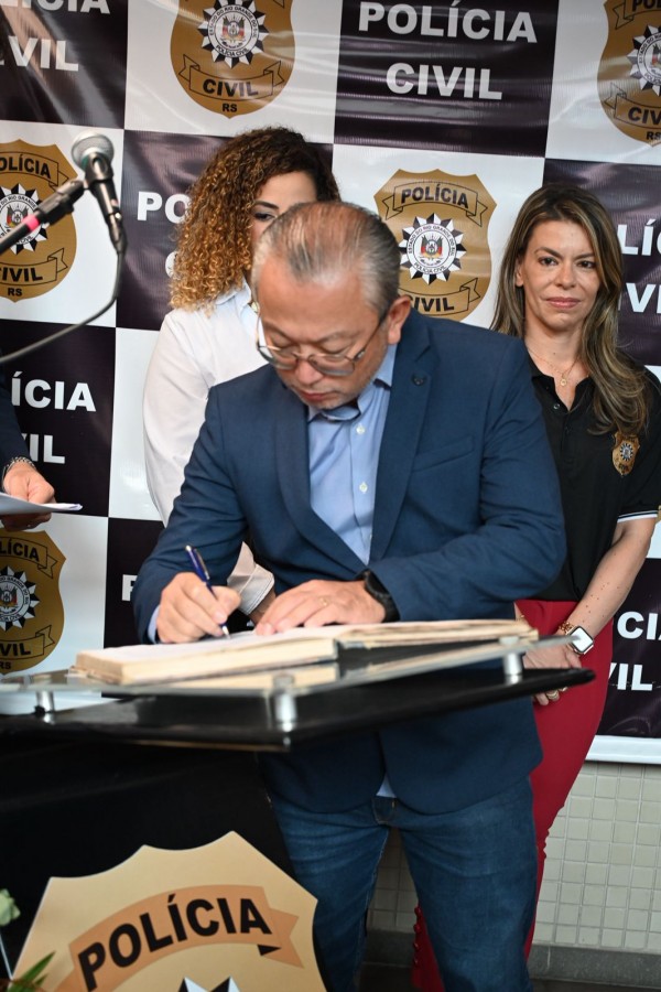 Esta é uma descrição da imagem para fins de acessibilidade:

A fotografia em plano médio e fechado registra o momento de uma assinatura oficial durante a inauguração de unidades policiais em Rio Grande.

Elementos Principais
O Ato da Assinatura: Em primeiro plano, o secretário adjunto da Segurança Pública, Mário Ikeda, veste um terno azul-marinho sobre uma camisa azul clara. Ele está levemente inclinado, usando óculos e concentrado enquanto assina um livro de atas ou documento oficial posicionado sobre um púlpito preto.

Autoridades e Observadores: * Atrás do secretário, à direita, uma mulher de cabelos longos e castanhos, vestindo a camiseta polo preta oficial da Polícia Civil e calça vermelha, observa o ato com uma expressão serena.

À esquerda, parcialmente visível atrás do secretário, está uma mulher de cabelos cacheados e volumosos.

Identificação Institucional: O fundo é composto por um painel (backdrop) xadrez em preto e branco, onde se alternam o brasão dourado da Polícia Civil do Rio Grande do Sul e a inscrição "POLÍCIA CIVIL" em letras brancas.

Detalhes e Ambiente
O Púlpito: A mesa de apoio possui uma base preta e um tampo de acrílico transparente fixado por suportes metálicos. Na parte frontal do púlpito, há um grande adesivo do distintivo da Polícia Civil.

Iluminação e Composição: A luz é clara e frontal, focando na ação das mãos do secretário. A composição destaca a formalidade do protocolo e a importância da medida para o fortalecimento da presença policial na região.