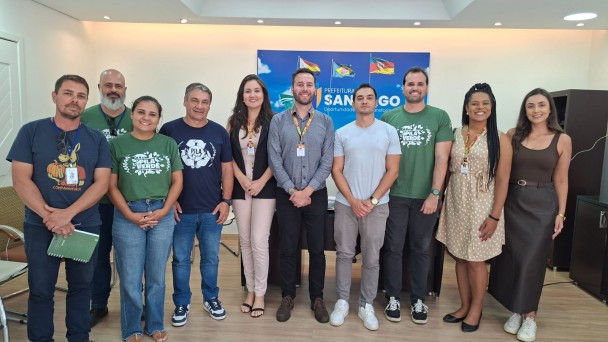 A imagem registra um grupo alinhado para uma foto institucional em uma sala com painel ao fundo contendo bandeiras estilizadas e o nome &ldquo;Prefeitura de Santiago&rdquo;. As pessoas est&atilde;o organizadas lado a lado, algumas usando crach&aacute;s e camisetas vinculadas ao projeto &ldquo;Pila Verde&rdquo;. 
