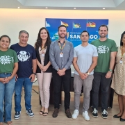 A imagem registra um grupo alinhado para uma foto institucional em uma sala com painel ao fundo contendo bandeiras estilizadas e o nome &ldquo;Prefeitura de Santiago&rdquo;. As pessoas est&atilde;o organizadas lado a lado, algumas usando crach&aacute;s e camisetas vinculadas ao projeto &ldquo;Pila Verde&rdquo;. 
