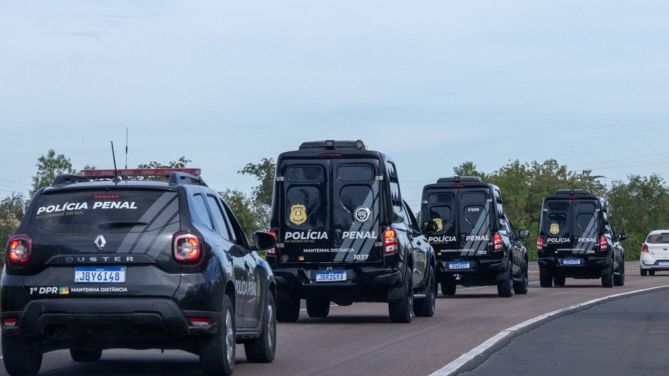 Esta &eacute; uma descri&ccedil;&atilde;o da imagem para fins de acessibilidade:

A fotografia registra um comboio de viaturas da Pol&iacute;cia Penal do Rio Grande do Sul em deslocamento por uma rodovia, ilustrando a marca de mais de 80 mil escoltas realizadas em 2025.

Elementos Principais
O Comboio: Quatro viaturas oficiais pretas est&atilde;o alinhadas em uma estrada de pista simples. As viaturas seguem em fila indiana, afastando-se da c&acirc;mera (vis&atilde;o traseira).

Tipos de Ve&iacute;culos:

Em primeiro plano, &agrave; esquerda, h&aacute; um utilit&aacute;rio modelo Renault Duster com o giroflex vermelho e azul ligado. Na parte superior do vidro traseiro, l&ecirc;-se "POL&Iacute;CIA PENAL" e, abaixo da placa, a identifica&ccedil;&atilde;o "1&ordm; DPR".

Logo &agrave; frente, seguem tr&ecirc;s furg&otilde;es de maior porte (estilo viaturas de transporte de presos ou escolta t&aacute;tica), tamb&eacute;m pretos, com grades nas janelas laterais e traseiras e a inscri&ccedil;&atilde;o "POL&Iacute;CIA PENAL" em destaque na lataria traseira.

Identifica&ccedil;&atilde;o: Todas as viaturas possuem o bras&atilde;o dourado da Pol&iacute;cia Penal e a orienta&ccedil;&atilde;o de seguran&ccedil;a "MANTENHA DIST&Acirc;NCIA" escrita em letras brancas.

Cen&aacute;rio e Ambiente
Rodovia: A estrada possui asfalto escuro com sinaliza&ccedil;&atilde;o de faixa branca cont&iacute;nua &agrave; direita. O comboio ocupa a faixa principal, enquanto ao fundo, &agrave; direita, um carro de passeio branco trafega no sentido oposto.

Paisagem: As margens da rodovia s&atilde;o compostas por vegeta&ccedil;&atilde;o rasteira e &aacute;rvores verdes sob um c&eacute;u claro e levemente nublado.

Composi&ccedil;&atilde;o: A foto utiliza uma perspectiva de profundidade, focando no alinhamento das viaturas, o que transmite uma sensa&ccedil;&atilde;o de organiza&ccedil;&atilde;o, seguran&ccedil;a e prontid&atilde;o operacional.