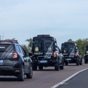 Esta &eacute; uma descri&ccedil;&atilde;o da imagem para fins de acessibilidade:

A fotografia registra um comboio de viaturas da Pol&iacute;cia Penal do Rio Grande do Sul em deslocamento por uma rodovia, ilustrando a marca de mais de 80 mil escoltas realizadas em 2025.

Elementos Principais
O Comboio: Quatro viaturas oficiais pretas est&atilde;o alinhadas em uma estrada de pista simples. As viaturas seguem em fila indiana, afastando-se da c&acirc;mera (vis&atilde;o traseira).

Tipos de Ve&iacute;culos:

Em primeiro plano, &agrave; esquerda, h&aacute; um utilit&aacute;rio modelo Renault Duster com o giroflex vermelho e azul ligado. Na parte superior do vidro traseiro, l&ecirc;-se "POL&Iacute;CIA PENAL" e, abaixo da placa, a identifica&ccedil;&atilde;o "1&ordm; DPR".

Logo &agrave; frente, seguem tr&ecirc;s furg&otilde;es de maior porte (estilo viaturas de transporte de presos ou escolta t&aacute;tica), tamb&eacute;m pretos, com grades nas janelas laterais e traseiras e a inscri&ccedil;&atilde;o "POL&Iacute;CIA PENAL" em destaque na lataria traseira.

Identifica&ccedil;&atilde;o: Todas as viaturas possuem o bras&atilde;o dourado da Pol&iacute;cia Penal e a orienta&ccedil;&atilde;o de seguran&ccedil;a "MANTENHA DIST&Acirc;NCIA" escrita em letras brancas.

Cen&aacute;rio e Ambiente
Rodovia: A estrada possui asfalto escuro com sinaliza&ccedil;&atilde;o de faixa branca cont&iacute;nua &agrave; direita. O comboio ocupa a faixa principal, enquanto ao fundo, &agrave; direita, um carro de passeio branco trafega no sentido oposto.

Paisagem: As margens da rodovia s&atilde;o compostas por vegeta&ccedil;&atilde;o rasteira e &aacute;rvores verdes sob um c&eacute;u claro e levemente nublado.

Composi&ccedil;&atilde;o: A foto utiliza uma perspectiva de profundidade, focando no alinhamento das viaturas, o que transmite uma sensa&ccedil;&atilde;o de organiza&ccedil;&atilde;o, seguran&ccedil;a e prontid&atilde;o operacional.