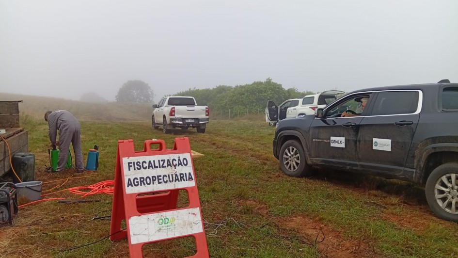 Imagem mostra uma estrada de terra e uma placa laranja onde est&aacute; escrito Fiscaliza&ccedil;&atilde;o Agropecu&aacute;ria. H&aacute; caminhonetes paradas atr&aacute;s e um t&eacute;cnico fazendo a fiscaliza&ccedil;&atilde;o. O dia est&aacute; com cerra&ccedil;&atilde;o baixa. 