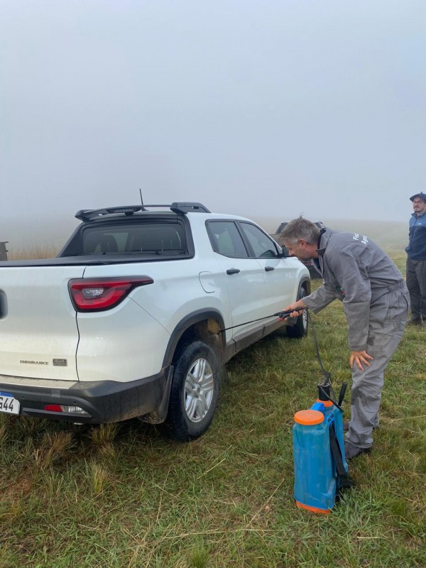Foto mostra técnico aplicando produtos nas rodas de uma caminhonete na beira de uma estrada de terra. O dia está com cerração baixa.