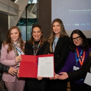 A fotografia registra um grupo de seis mulheres durante um evento internacional na ONU, celebrando a assinatura de um termo de coopera&ccedil;&atilde;o entre os estados do COSUD (Cons&oacute;rcio de Integra&ccedil;&atilde;o Sul e Sudeste) para o fortalecimento de pol&iacute;ticas para mulheres.

Elementos Principais
O Grupo: Seis mulheres est&atilde;o posicionadas lado a lado, em plano m&eacute;dio, sorrindo para a c&acirc;mera. Elas possuem diferentes perfis e estilos, vestindo blazers, jaquetas e roupas sociais em tons de preto, rosa e roxo. Todas utilizam crach&aacute;s com cord&otilde;es coloridos t&iacute;picos de confer&ecirc;ncias internacionais.

O Documento: No centro, tr&ecirc;s das mulheres seguram juntas uma pasta de couro vermelha aberta, exibindo um documento oficial de uma p&aacute;gina com o bras&atilde;o do Estado do Rio Grande do Sul no topo.

A Proje&ccedil;&atilde;o: Ao fundo, &agrave; direita, uma tela exibe o t&iacute;tulo do evento: "F&oacute;rum Sobre Masculinidades - RESPOSTAS REGIONAIS PARA PROBLEMAS GLOBAIS". Abaixo do t&iacute;tulo, h&aacute; logotipos institucionais, incluindo o da ONU Mulheres e do Governo do Brasil.

Detalhes e Ambiente
Cen&aacute;rio: O ambiente &eacute; sofisticado e moderno. &Agrave; esquerda, destaca-se uma estrutura arquitet&ocirc;nica com janelas de vidro que mostram o reflexo de pr&eacute;dios urbanos, sugerindo a sede da ONU em Nova York.

Composi&ccedil;&atilde;o: A imagem foca na uni&atilde;o das m&atilde;os segurando o documento vermelho, simbolizando a coopera&ccedil;&atilde;o e o compromisso firmado entre as gestoras dos diferentes estados.

Ilumina&ccedil;&atilde;o: A luz &eacute; suave e bem distribu&iacute;da, ressaltando os detalhes dos rostos e a formalidade do encontro.