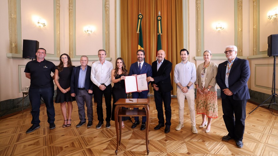 A foto mostra um grupo alinhado para registro oficial em um sal&atilde;o hist&oacute;rico, com o governador ao centro, que segura um documento aberto sobre uma mesa. Ao fundo, duas bandeiras est&atilde;o posicionadas diante da cortina dourada.