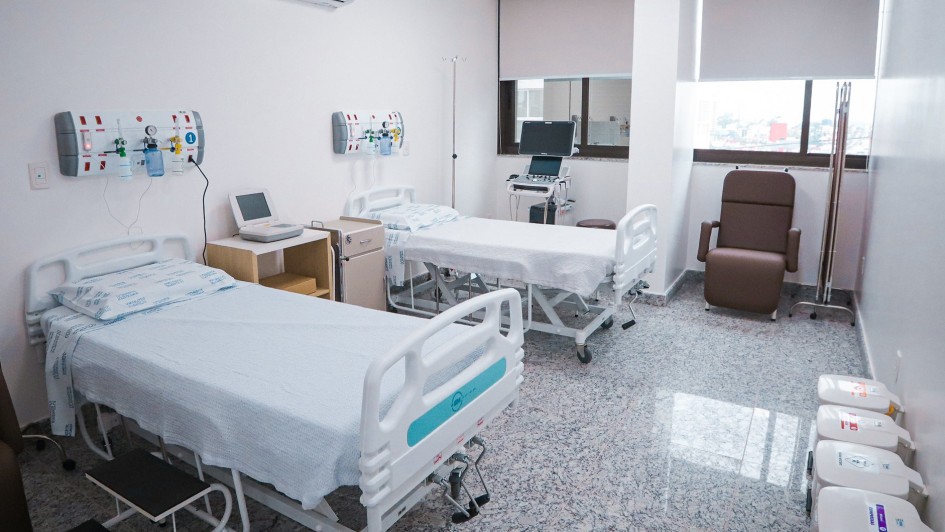 Esta &eacute; uma descri&ccedil;&atilde;o da imagem para fins de acessibilidade:

A fotografia registra o interior de um quarto hospitalar moderno e equipado no Hospital de Cl&iacute;nicas de Passo Fundo, apresentando um ambiente limpo e organizado.

Elementos Principais
Leitos Hospitalares: O quarto disp&otilde;e de dois leitos com grades de prote&ccedil;&atilde;o brancas e colchas claras. O leito em primeiro plano possui uma escadinha de dois degraus pretos ao lado.

Equipamentos M&eacute;dicos: * Acima de cada cabeceira, h&aacute; pain&eacute;is de gases medicinais (com flux&ocirc;metros e sa&iacute;das de oxig&ecirc;nio).

Entre as camas, encontra-se um monitor m&eacute;dico sobre um m&oacute;vel de apoio e um suporte para soro.

Ao fundo, pr&oacute;ximo &agrave; janela, h&aacute; um equipamento de diagn&oacute;stico com tela (possivelmente um ultrassom ou monitor avan&ccedil;ado).

Mobili&aacute;rio de Apoio: &Agrave; direita, junto &agrave; janela, h&aacute; uma poltrona de descanso reclin&aacute;vel em tom marrom. No canto inferior direito, est&atilde;o alinhados quatro cestos de lixo brancos com tampas identificadas por cores para descarte seletivo de res&iacute;duos hospitalares.

Detalhes e Ambiente
Infraestrutura: O quarto possui piso de granito cinza polido, paredes brancas e um aparelho de ar-condicionado split instalado no alto da parede esquerda.

Ilumina&ccedil;&atilde;o e Ventila&ccedil;&atilde;o: Uma ampla janela ao fundo, com persianas integradas, permite a entrada de luz natural e oferece uma vista parcial de pr&eacute;dios da cidade. A ilumina&ccedil;&atilde;o artificial &eacute; feita por pain&eacute;is de LED embutidos no teto.

Composi&ccedil;&atilde;o: A imagem &eacute; capturada de um &acirc;ngulo que privilegia a profundidade do quarto, destacando a prontid&atilde;o da estrutura para receber pacientes.