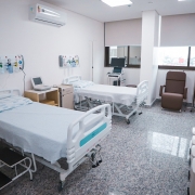 Esta &eacute; uma descri&ccedil;&atilde;o da imagem para fins de acessibilidade:

A fotografia registra o interior de um quarto hospitalar moderno e equipado no Hospital de Cl&iacute;nicas de Passo Fundo, apresentando um ambiente limpo e organizado.

Elementos Principais
Leitos Hospitalares: O quarto disp&otilde;e de dois leitos com grades de prote&ccedil;&atilde;o brancas e colchas claras. O leito em primeiro plano possui uma escadinha de dois degraus pretos ao lado.

Equipamentos M&eacute;dicos: * Acima de cada cabeceira, h&aacute; pain&eacute;is de gases medicinais (com flux&ocirc;metros e sa&iacute;das de oxig&ecirc;nio).

Entre as camas, encontra-se um monitor m&eacute;dico sobre um m&oacute;vel de apoio e um suporte para soro.

Ao fundo, pr&oacute;ximo &agrave; janela, h&aacute; um equipamento de diagn&oacute;stico com tela (possivelmente um ultrassom ou monitor avan&ccedil;ado).

Mobili&aacute;rio de Apoio: &Agrave; direita, junto &agrave; janela, h&aacute; uma poltrona de descanso reclin&aacute;vel em tom marrom. No canto inferior direito, est&atilde;o alinhados quatro cestos de lixo brancos com tampas identificadas por cores para descarte seletivo de res&iacute;duos hospitalares.

Detalhes e Ambiente
Infraestrutura: O quarto possui piso de granito cinza polido, paredes brancas e um aparelho de ar-condicionado split instalado no alto da parede esquerda.

Ilumina&ccedil;&atilde;o e Ventila&ccedil;&atilde;o: Uma ampla janela ao fundo, com persianas integradas, permite a entrada de luz natural e oferece uma vista parcial de pr&eacute;dios da cidade. A ilumina&ccedil;&atilde;o artificial &eacute; feita por pain&eacute;is de LED embutidos no teto.

Composi&ccedil;&atilde;o: A imagem &eacute; capturada de um &acirc;ngulo que privilegia a profundidade do quarto, destacando a prontid&atilde;o da estrutura para receber pacientes.
