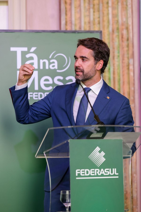 A imagem mostra o governador falando ao microfone em um púlpito transparente com a marca Federasul. Ao fundo, há um painel verde com a identidade visual do evento Tá na Mesa – Federasul.