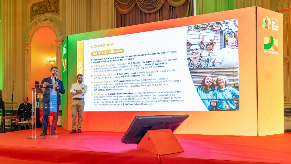 A foto registra um palco amplo, onde o governador e o vice-governador est&atilde;o posicionados &agrave; frente de um tel&atilde;o de grandes dimens&otilde;es. O painel exibe conte&uacute;do sobre Economia, com dados, valores e imagens ilustrativas. 