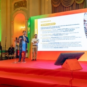 A foto registra um palco amplo, onde o governador e o vice-governador est&atilde;o posicionados &agrave; frente de um tel&atilde;o de grandes dimens&otilde;es. O painel exibe conte&uacute;do sobre Economia, com dados, valores e imagens ilustrativas. 