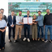 Um grupo de autoridades posa em frente a um painel do programa RS Seguro. No centro, o governador Eduardo Leite segura um documento aberto ao lado de outros homens e uma mulher. &Agrave; esquerda, uma mulher de &oacute;culos est&aacute; em primeiro plano; ao fundo, um homem fala ao microfone.