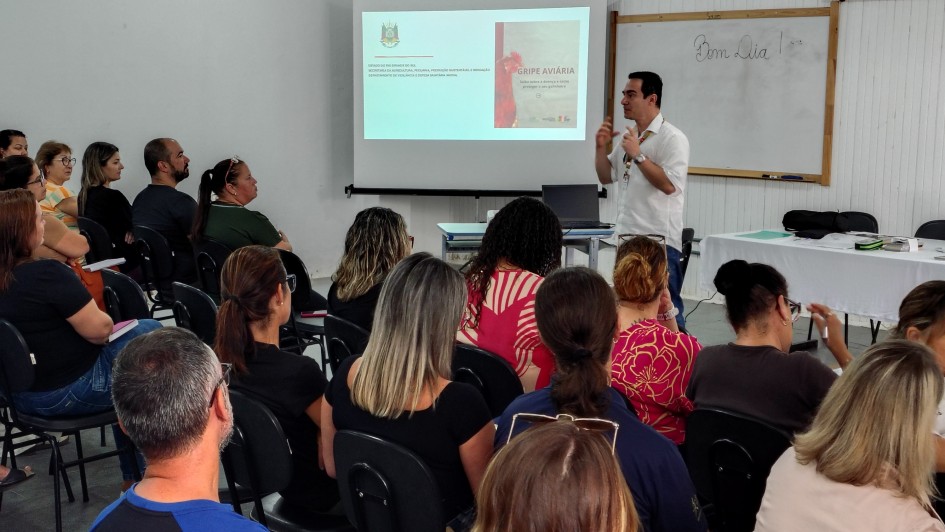 A imagem mostra uma sala cheia de pessoas sentadas assistindo a uma palestra. Um homem est&aacute; em p&eacute;, falando com o p&uacute;blico. Ao fundo, h&aacute; um projeto de slides com a apresenta&ccedil;&atilde;o, uma mesa e um quadro branco.