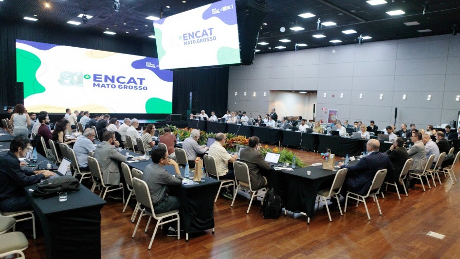 A imagem mostra um grande audit&oacute;rio preparado para um encontro institucional, com mesas retangulares dispostas em formato de &ldquo;U&rdquo; e ocupadas por participantes que utilizam notebooks, pastas e garrafas de &aacute;gua. No fundo do sal&atilde;o, um tel&atilde;o amplo exibe a identidade visual do evento: &ldquo;ENCAT Mato Grosso&rdquo;, com cores verde, azul e amarelo.