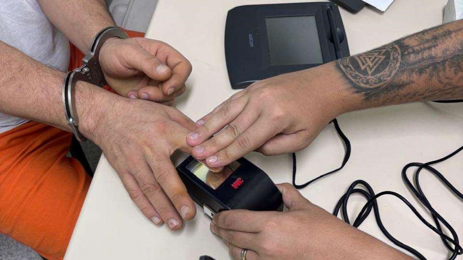 Foto mostra o detalhe da m&atilde;o de um detento colocando o dedo no leitor digital com ajuda do servidor. Ele est&aacute; algemado e aparece parte da cal&ccedil;a laranja do uniforme. 