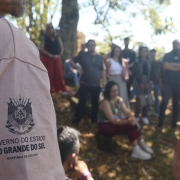 A imagem mostra, à direita, o brasão do Estado do Rio Grande do Sul acima da inscrição "Secretaria da Cultura" impresso na manga de uma camiseta. Ao fundo, aparece um grupo de turistas em desfoque em uma área sombreada.