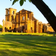 A imagem mostra a fachada das Ruínas de São Miguel das Missões ensolarada emoldurada por troncos de árvore.