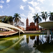 A imagem mostra uma vista da Praça Pinheiro Machado, de Santo Ângelo, com o lago central, chafariz desligado, e a catedral angelopolitana ao fundo. Às margens da imagem, nota-se palmeiras altas contornando a praça.