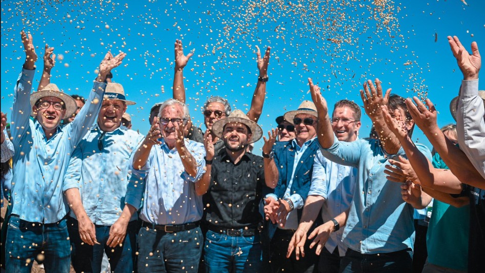Grupo de cerca de dez pessoas joga gr&atilde;os de soja para o alto em comemora&ccedil;&atilde;o, sob c&eacute;u azul intenso. V&aacute;rios usam chap&eacute;us de palha e camisas claras. No centro, destaca-se um homem de preto. A cena celebra a abertura da colheita da soja.