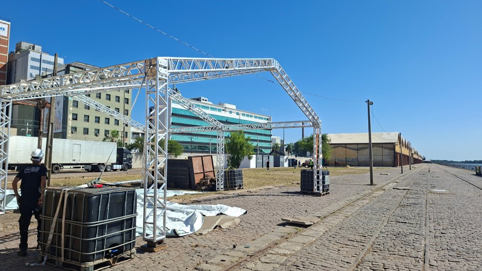 A fotografia registra os preparativos estruturais no Cais Mau&aacute;, em Porto Alegre, para a realiza&ccedil;&atilde;o do evento de inova&ccedil;&atilde;o South Summit Brazil 2026.

Elementos Principais
Estrutura Met&aacute;lica: Em primeiro plano, destaca-se a arma&ccedil;&atilde;o de uma grande tenda ou pavilh&atilde;o feita de treli&ccedil;as de alum&iacute;nio (box truss). A estrutura est&aacute; em fase de montagem, posicionada sobre o cal&ccedil;amento de paralelep&iacute;pedos.

Base de Sustenta&ccedil;&atilde;o: Os pilares da estrutura est&atilde;o fixados e estabilizados por grandes reservat&oacute;rios pl&aacute;sticos pretos (IBCs) cheios de &aacute;gua, que servem como contrapesos.

Trabalhador: &Agrave; esquerda, um oper&aacute;rio de costas, vestindo camiseta preta e capacete de seguran&ccedil;a branco, observa a montagem.

Cen&aacute;rio e Ambiente
Cais Mau&aacute;: &Agrave; direita, estendem-se os trilhos hist&oacute;ricos de trem e a linha de armaz&eacute;ns amarelos caracter&iacute;sticos do porto, que margeiam o Lago Gua&iacute;ba (vis&iacute;vel ao fundo, &agrave; extrema direita).

Entorno Urbano: Ao fundo, o cen&aacute;rio &eacute; composto por pr&eacute;dios comerciais, incluindo um edif&iacute;cio com fachada de vidro verde e outros de arquitetura mais tradicional. Um caminh&atilde;o branco est&aacute; estacionado na &aacute;rea de log&iacute;stica &agrave; esquerda.

Condi&ccedil;&otilde;es Clim&aacute;ticas: O dia est&aacute; ensolarado, com c&eacute;u azul sem nuvens, o que proporciona uma ilumina&ccedil;&atilde;o clara e sombras n&iacute;tidas no ch&atilde;o.

Composi&ccedil;&atilde;o e Contexto
A imagem utiliza uma perspectiva que valoriza a extens&atilde;o do Cais Mau&aacute;, conectando a infraestrutura tempor&aacute;ria do evento ao patrim&ocirc;nio hist&oacute;rico da cidade.

O registro enfatiza a fase de "ajustes finais" e montagem log&iacute;stica para receber o p&uacute;blico do festival de inova&ccedil;&atilde;o.