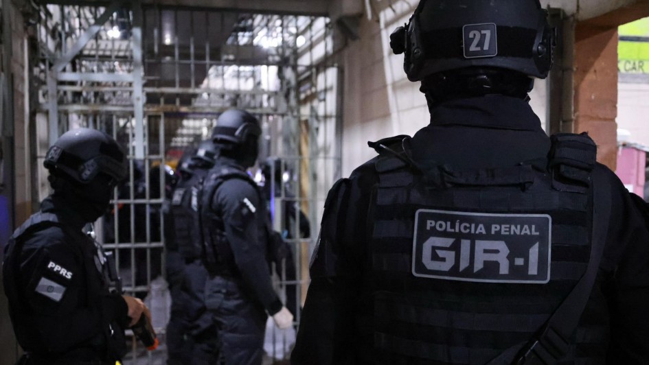 Foto mostra policiais penais, de costa, e com uniformes pretos e capacetes, no corredor do Complexo Prisional de Canoas, ao fundo aparecem grades no meio do corredor. 