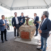 A fotografia registra o ato solene de lan&ccedil;amento da pedra fundamental do resort Club Med Gramado, com a participa&ccedil;&atilde;o do governador Eduardo Leite e executivos do empreendimento.

Elementos Principais
O Monumento: No centro da imagem, sobre uma base de concreto, repousa uma grande rocha natural (pedra fundamental). Nela, est&aacute; fixada uma placa met&aacute;lica avermelhada com inscri&ccedil;&otilde;es comemorativas. Sobre a pedra, h&aacute; uma escultura de metal composta por dois arcos que se cruzam.

O Governador: Eduardo Leite, vestindo um blazer azul-marinho sobre uma camiseta branca e cal&ccedil;a cinza, est&aacute; posicionado atr&aacute;s da pedra. Ele segura um pequeno objeto cil&iacute;ndrico (provavelmente uma c&aacute;psula do tempo) que est&aacute; sendo depositado no topo do monumento.

As Autoridades e Convidados: Cerca de oito homens e uma mulher cercam o monumento sob uma tenda. Eles vestem trajes esporte fino (blazers e camisas sociais). Alguns seguram ta&ccedil;as com espumante, celebrando o momento com sorrisos.

Cen&aacute;rio e Ambiente
Local: O evento ocorre em uma &aacute;rea elevada e aberta. Ao fundo, avista-se uma vasta paisagem natural caracter&iacute;stica da Serra Ga&uacute;cha, com muitas copas de &aacute;rvores (arauc&aacute;rias) e o horizonte sob um c&eacute;u azul com nuvens esparsas.

Estrutura: O ato &eacute; protegido por uma tenda de lona branca. O ch&atilde;o &eacute; coberto por brita cinza.

Composi&ccedil;&atilde;o: A foto captura o grupo por inteiro, destacando a integra&ccedil;&atilde;o entre o investimento privado e o cen&aacute;rio natural da regi&atilde;o.