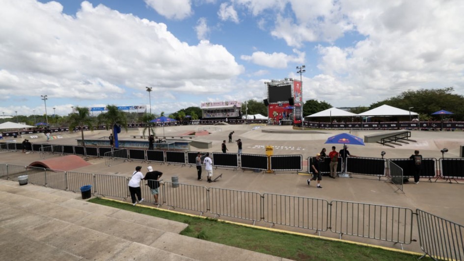 A fotografia registra a estrutura da maior pista de skate da Am&eacute;rica Latina, na Orla do Gua&iacute;ba, preparada para a abertura do STU National 2026 em Porto Alegre.

Elementos Principais
A Pista (Skatepark): Em plano m&eacute;dio, v&ecirc;-se a vasta &aacute;rea de concreto liso composta por diferentes obst&aacute;culos de modalidades como Street e Park, incluindo rampas, corrim&atilde;os e v&atilde;os t&eacute;cnicos. O asfalto exibe a inscri&ccedil;&atilde;o "STU NATIONAL PORTO ALEGRE" centralizada no piso.

Estrutura de Evento: Ao fundo, destacam-se grandes tendas brancas, arquibancadas met&aacute;licas laterais e uma torre central com um tel&atilde;o de LED e o logotipo do evento em fundo vermelho. Diversos ombrelones (guarda-s&oacute;is) azuis da marca Red Bull est&atilde;o espalhados pela &aacute;rea t&eacute;cnica.

Pessoas e Movimenta&ccedil;&atilde;o: Algumas poucas pessoas, entre skatistas e equipe t&eacute;cnica, circulam pela pista. Em primeiro plano, dois jovens de camisetas brancas e bon&eacute;s observam a pista apoiados em uma grade met&aacute;lica de conten&ccedil;&atilde;o.

Detalhes e Ambiente
Cen&aacute;rio: A imagem &eacute; capturada a partir de um ponto elevado (provavelmente o topo da arquibancada de concreto). O horizonte &eacute; marcado por &aacute;rvores verdes e o c&eacute;u est&aacute; azul, densamente povoado por nuvens brancas e volumosas.

Ilumina&ccedil;&atilde;o: A luz solar &eacute; difusa devido &agrave;s nuvens, criando uma ilumina&ccedil;&atilde;o clara e uniforme sobre todo o complexo esportivo.
