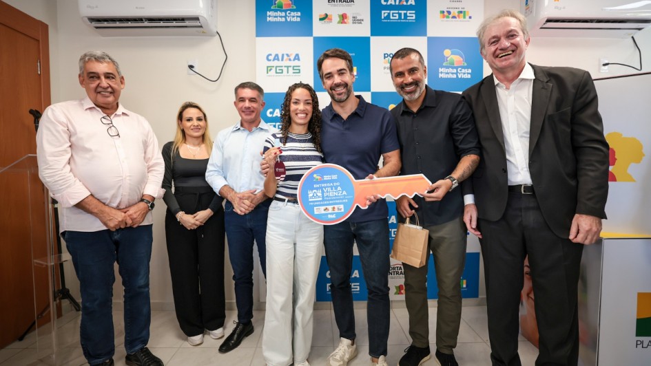 A imagem mostra um grupo reunido em um ambiente interno durante a entrega simb&oacute;lica de moradias. No centro, v&aacute;rias pessoas seguram uma chave cenogr&aacute;fica gigante, identificada com o texto &ldquo;Entrega do Residencial Villa Pienza &ndash; PAC / Minha Casa Minha Vida&rdquo;. O fundo &eacute; composto por um painel com logomarcas da Caixa, FGTS, Minha Casa Minha Vida e Governo Federal.