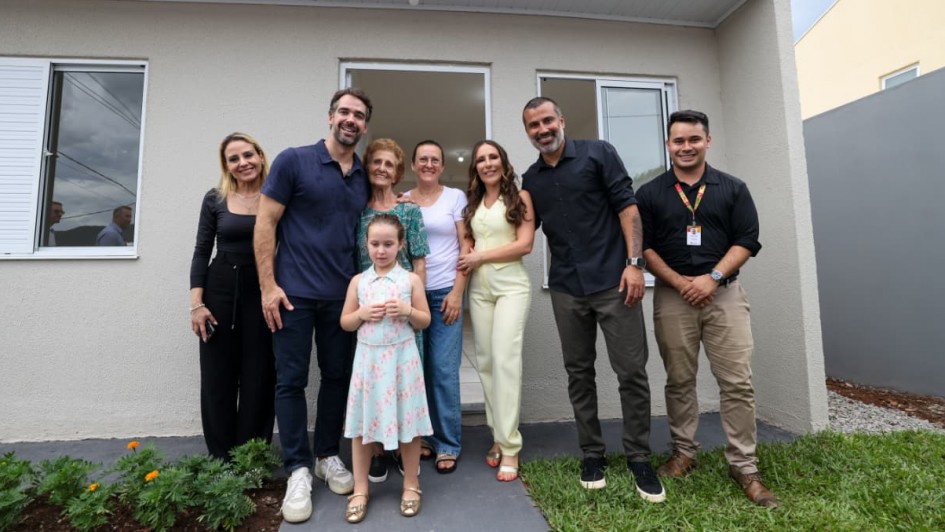 Grupo posicionado em frente a uma casa t&eacute;rrea rec&eacute;m-constru&iacute;da, com fachada clara, porta branca e janela lateral. As pessoas, incluindo o governador, est&atilde;o alinhadas para foto sobre uma &aacute;rea gramada e cal&ccedil;ada de acesso.