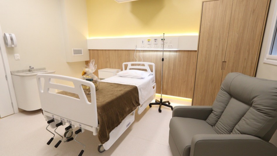 Imagem de um quarto hospitalar com cama el&eacute;trica, poltrona reclin&aacute;vel, suporte para soro, arm&aacute;rio e pia. O ambiente &eacute; moderno, bem iluminado e organizado, com detalhes em madeira e luz indireta na parede.
