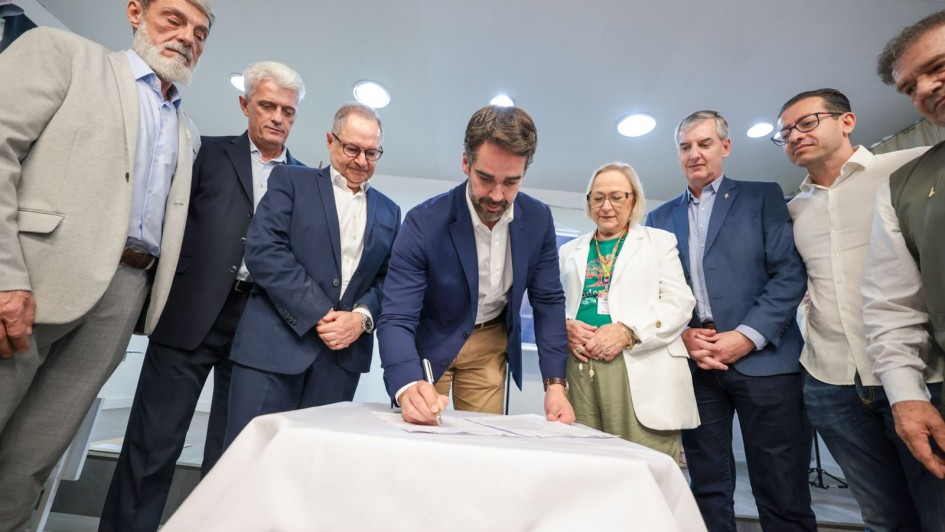 Grupo de pessoas em volta de uma mesa; Leite, ao centro, assina um documento enquanto os demais observam.