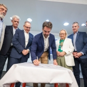 Grupo de pessoas em volta de uma mesa; Leite, ao centro, assina um documento enquanto os demais observam.