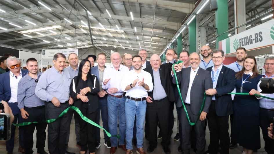A foto registra uma cerim&ocirc;nia de inaugura&ccedil;&atilde;o em pavilh&atilde;o coberto. Um grupo grande est&aacute; alinhado segurando uma fita verde para o corte simb&oacute;lico, com o vice-governador ao centro. O local possui estandes ao fundo e ilumina&ccedil;&atilde;o t&iacute;pica de galp&atilde;o de eventos, com estrutura met&aacute;lica e corredores amplos.