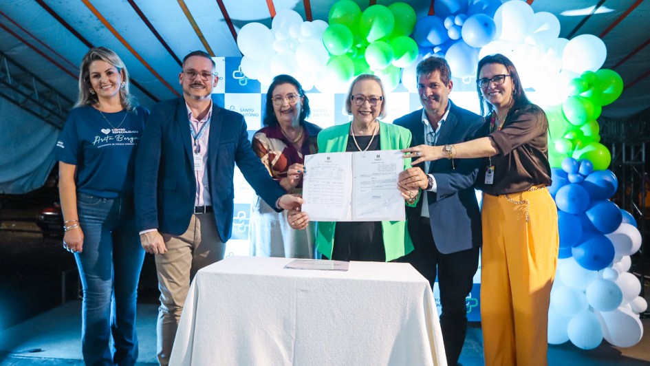 Foto posada na solenidade, com seis pessoas de p&eacute;, exibindo para a c&acirc;mera um documento assinado. Elas est&atilde;o atr&aacute;s de uma mesa e na frente de enfeites de bal&atilde;o. Todas sorriem.